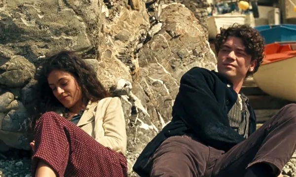 A still from the film Weightless. A young woman and man with Mediterranean features lounge against a rocky harbour wall.