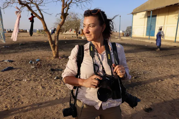 The photographer Lynsey Addario, a white woman walking past a low building, carrying her camera, with a bare tree behind her.