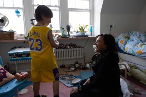 The photographer Lynsey Addario, a blond white woman kneeling on a bedroom floor beside a young white boy who is wearing a yellow football strip marked 'James 52'.