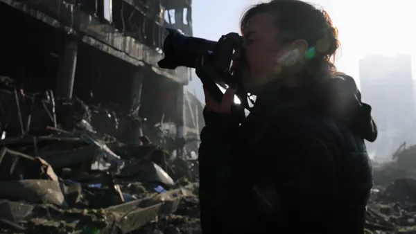 The photographer Lynsey Addario, a blond white woman pointing a large camera at a ruined building.