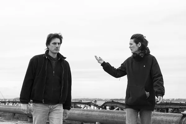A black and white photograph of a white man and woman walking along a waterfront. She is wearing headphones around her neck and gesticulating at him.