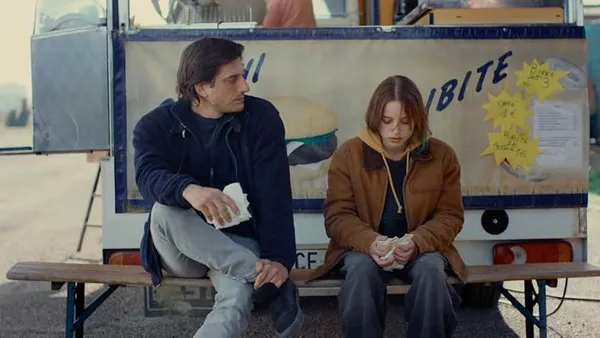 A still from the film Paternal Leave. A white man and girl sit on a bench beside a burger van. He is looking at her in a friendly way; she is looking down at her food unhappily.