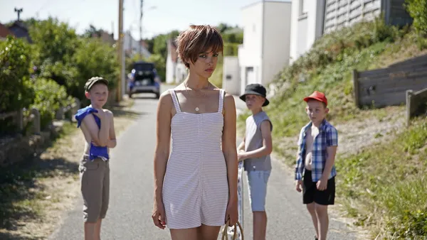 Lyna Khoudri. A young Black woman in summer clothing walks down a suburban street, with three young white boys behind her.
