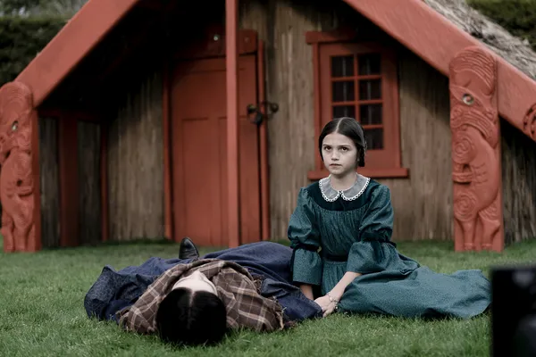 A still from the film Mārama. A Māori girl sits by the side of a Māori woman who is lying on her back in front of a wharenui., or ancestral hall