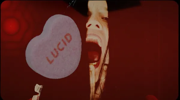 A still from the film Lucid. Heart-shaped candy with the word 'lucid' printed on it floats at one side of a red screen. A young white woman's screaming face can be seen through a rip in the redness.