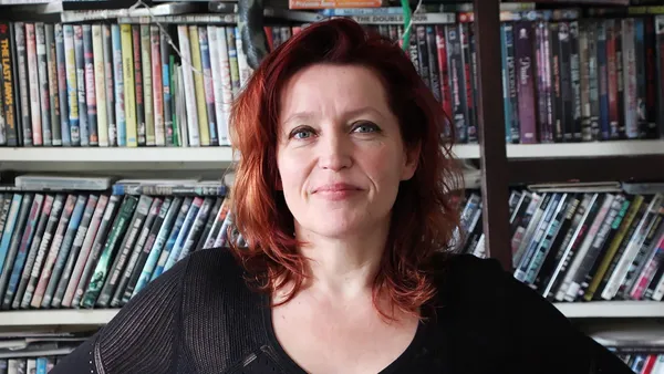 The filmmaker Tallulah H Schwab. A white woman with thick red hair, wearing a black top and standing in front of shelves full of DVDs cases.