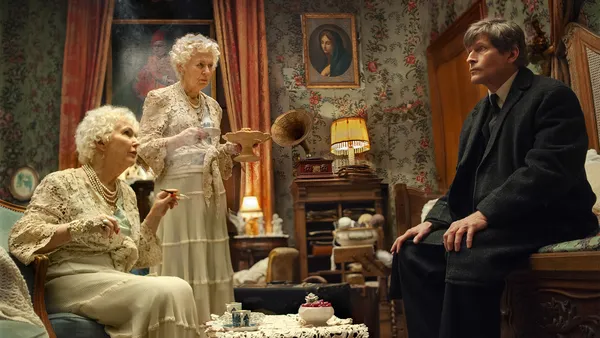A still from the film Mr. K. In a cluttered room with floral wallpaper, two white-haired elderly white women in white lacy clothes take tea with a middle-aged man in a dark suit.