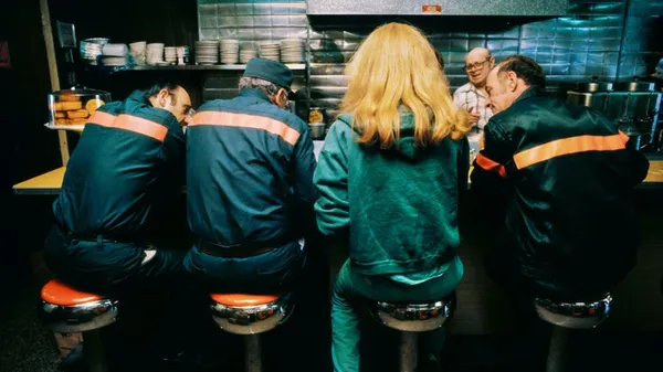 A still from the film Maintenance Artist. Three older man in work uniforms with fluorescent stripes sit at a food counter. With them is a figure in ble-green clothes with a large bob of yellow hair.