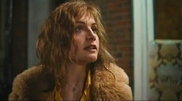 A young white woman with unkempt golden hair, a yellow top and a jacket trimmed with tawny fur, looking upwards.