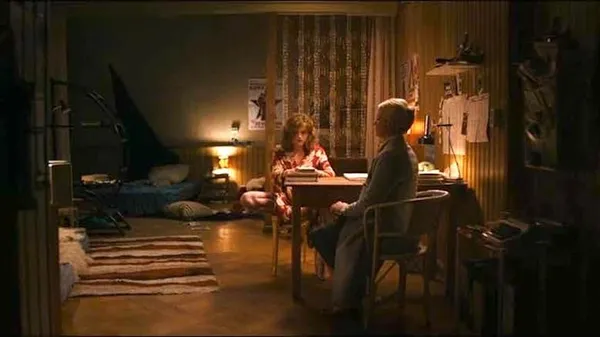 In an untidy room with mattresses on the floor, a young white woman site at a table with one leg folded under her, whilst a stiff-backed older white woman sits opposite her.
