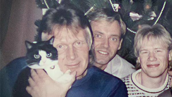 Three fair haired white men stand in front of a Christmas tree. One of them is hugging a black and white kitten.