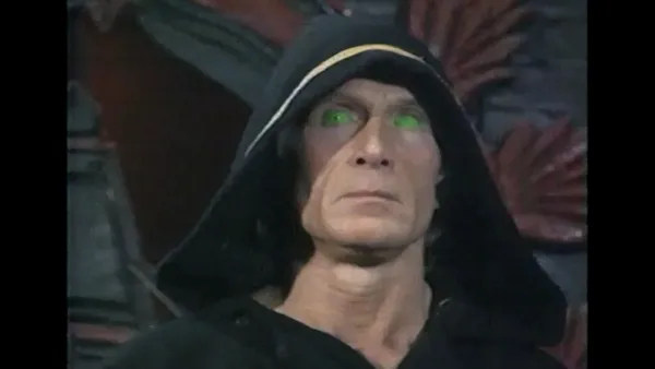 A still from the film The Eye Of Satan. A middle-aged white man with glowing green eyes, wearing a dark cowl.
