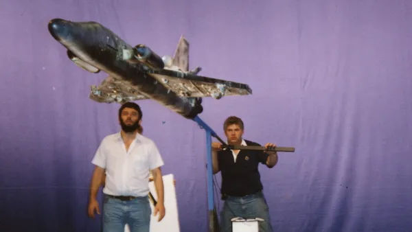 Two men manoeuvre a model spaceship on a metal frame, against a bluescreen background.