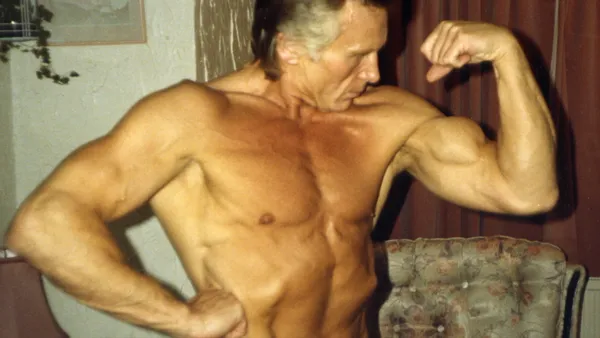 The filmmaker Cliff Twemlow. A shirtless, muscular middle-aged white man adopts a bodybuilding pose in his living room.