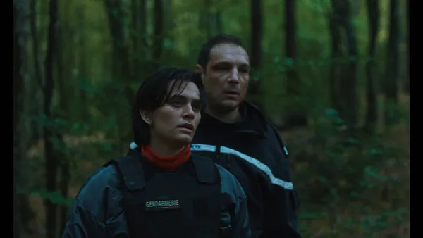 A still from the film Too Many Beasts. A white man with a bruised face follows a white woman who is wearing a bulletproof vest marked 'gendarmerie' into the woods.