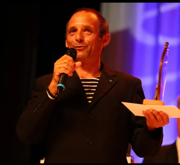 The filmmaker Mark Jenkins. A white man wearing a black and white horizontally striped top under a black jacket holds an award in his left hand and a microphone in his right.