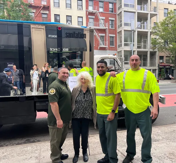 A blond middle-aged white woman stands with three sanitation workers, one Black and two white, beside a mirrored truck in an urban street.
