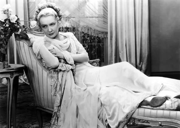 A black and white still from the film Becky Sharp. A white woman with elaborately coiffed blond hair and multi-layered lacy clothing lounges on a chaise longue in front of a window.