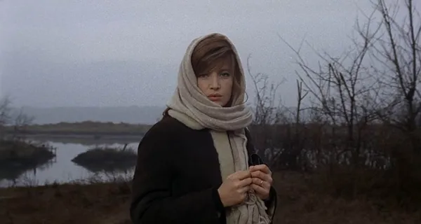 A still from the film Red Desert. A white woman wearing a warm, dark coat, with a pale grey scarf wrapped around her head, stands among leafless trees in marshland.