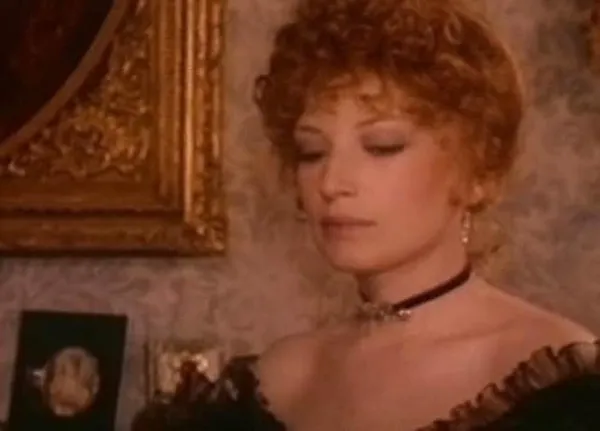 A still from the film The Mystery Of Oberwald. An elegantly dressed white woman stands against a wall with expensive wallpaper and the corner of a gilded plaster picture frame.
