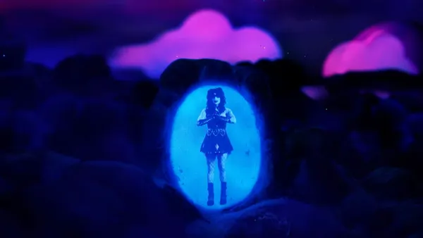 A still from the film The People's Joker. A blue and white image of a young white woman wearing a short dress, fishnets, big boots, gauntlets and clown make-up appears in an illuminated oval in a pile of rocks under a dark sky with flashes of pink.