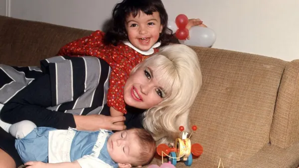 A smiling white woman with big blond hair lies on a brown sofa with a sleepy baby and a small girl who looks like the happiest child in the world.