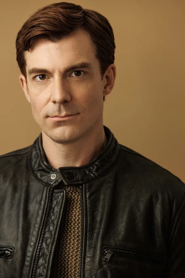 A white man with neat dark brown hair, a black jacket and a brown jumper.