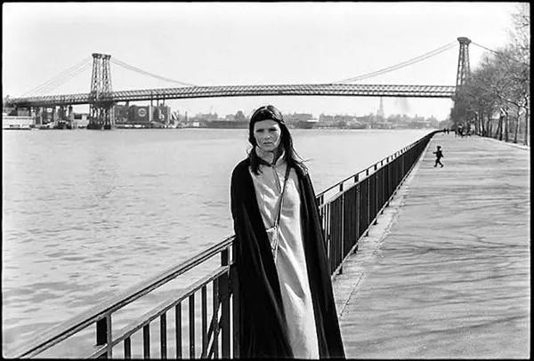 In black and white, a dark-haired white woman wearing a long, pale dress and soft black velvet cloak stands beside the railings on a riverside promenade, a large bridge visible behind her.