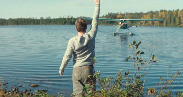 A still from the film Who By Fire. A white man stands by a lake, waving as a seaplane comes in to land.