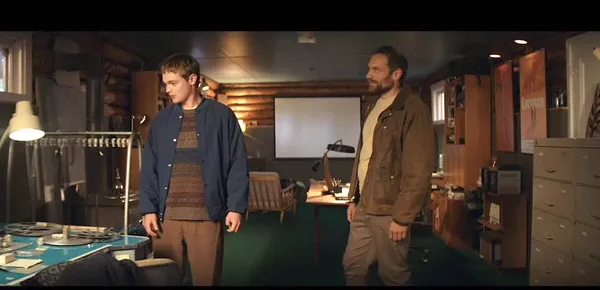 A still from the film Who By Fire. Two white men stand in a small room with a window behind them.
