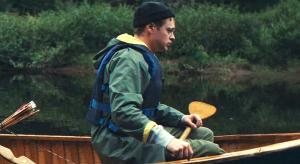 A still from the film Who By Fire. A young white man rows a boat along a river whose banks are thick with trees.
