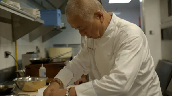 A still from the film Nobu. A chef with light brown skin and a shaved head is hard at work in a kitchen.
