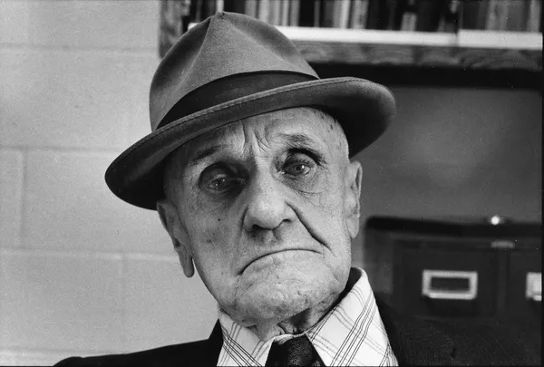 The face of a melancholy-looking elderly white man who is wearing a hat.
