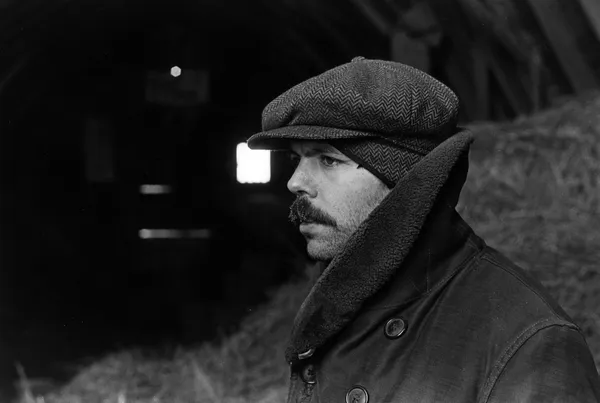 A lack and white head and shoulders shot of a middle aged, moustachioed white man wearing a flat cap and with his jacket collar pulled up, standing in front of a barn.