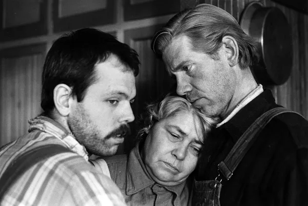 A black and white image of two men standing one on either side of an older woman. She is resting her head against the shoulder of one of them, and looks sad; they look sympathetic.