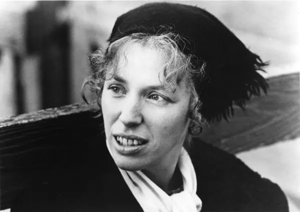 A black and white image of a toothy blond white woman wearing a black headscarf and leaning against a fence.