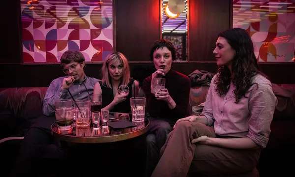 A still from the film Odyssey. Three white woman and a white man sit around a table in a nightclub. It is piled with glasses.