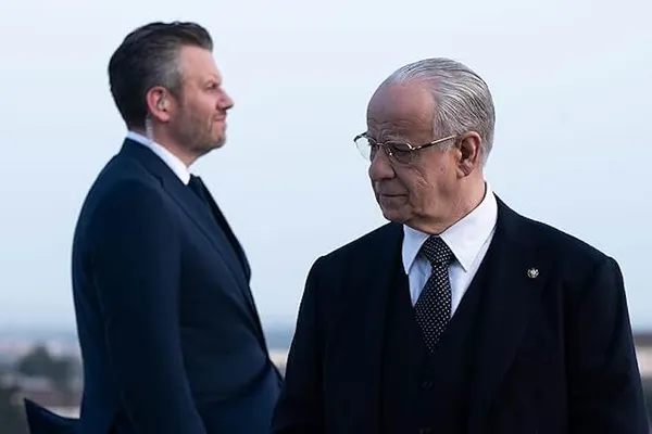 A still from the film La Grazia. An older white man in an expensive suit glances curiously at the large white male security guard standing behind him.