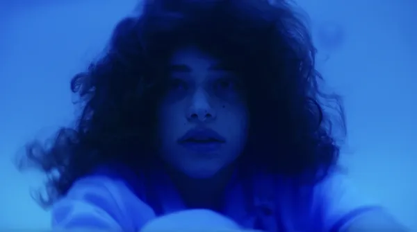 A still from the film Pools. A young woman with light coloured skin, dark curly hair and a nose ring sits at the bottom of a swimming pool.