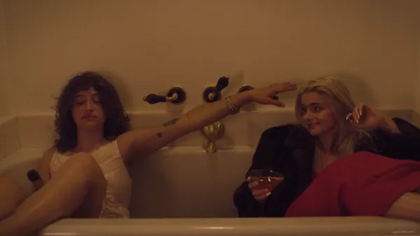 A still from the film Pools. Two young white women, one brunette and one blond, lounge in an empty bathtub drinking champagne, their feet hanging over the side.