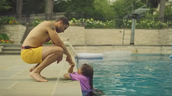 A still from the film Pools. A tanned, muscular young man in long yellow shorts squats beside an outdoor swimming pool, holding the hand of a young white woman in a pink shirt who is in the water.