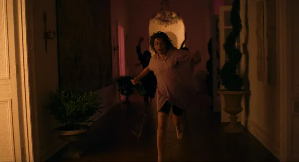 A still from the film Pools. A young white woman runs through the hallway of an expensive home, looking excited. There are other, indistinct figures behind her.