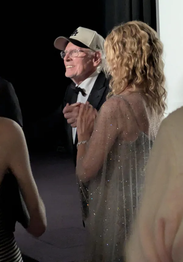 The actors Bruce Dern and Laura Dern. A tall blond white woman in a silver dress, with her back to the camera, supports an older, bespectacled white man who looks a little fragile, but alert.