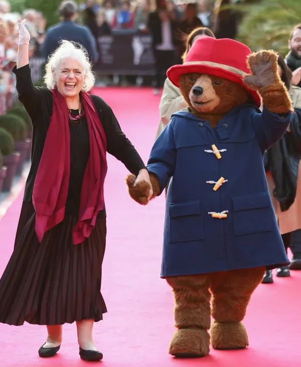 An older white woman with white hair, black clothing and a long red scarf walks along a red carpet hand in hand with Paddington Bear, who is wearing his trademark blue duffle coat and red hat. Both are waving.
