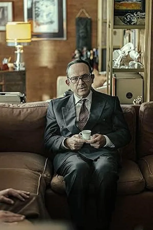 A dtill from the film The Great Ambition. A bespectacled middle-aged white man in a dark suit sits on a brown leather sofa, holding a tiny teacup and saucer, in a room that looks like a museum.