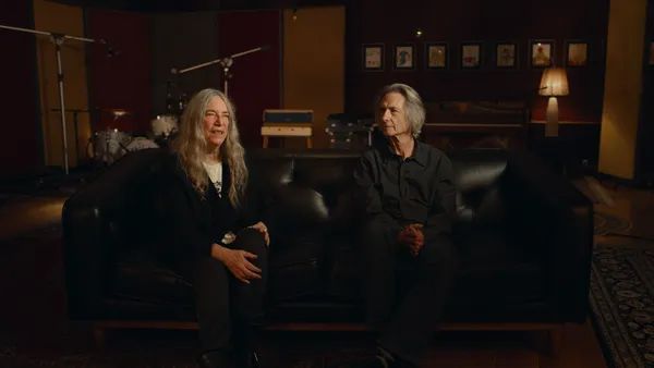 An older white woman and man, both wearing black trousers and shirts, sit on a black leather sofa in a room which has Afghan rugs on the floor and a drumkit in one corner.