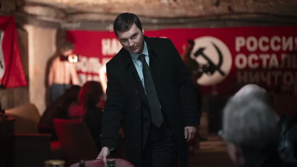A still from the film The Wizard Of The Kremlin. A young white man takes a seat in a bar which has a hammer and sickle banner hanging on one wall.