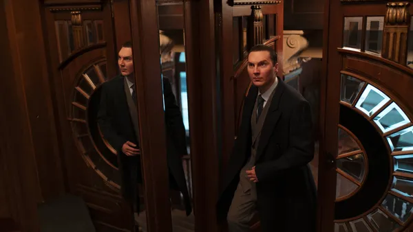 A still from the film The Wizard Of The Kremlin. A young white man is reflected in full length mirrors as he enters a wood-panelled room.