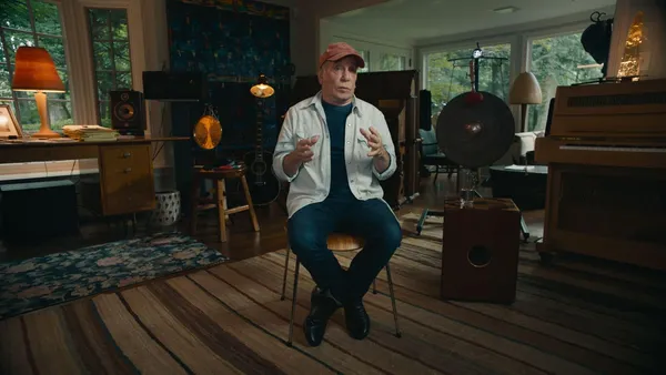 An older white man sits on a small chair in the centre of a large room full of antique furniture and music instruments, with a lush garden visible through two windows on adjacent walls.