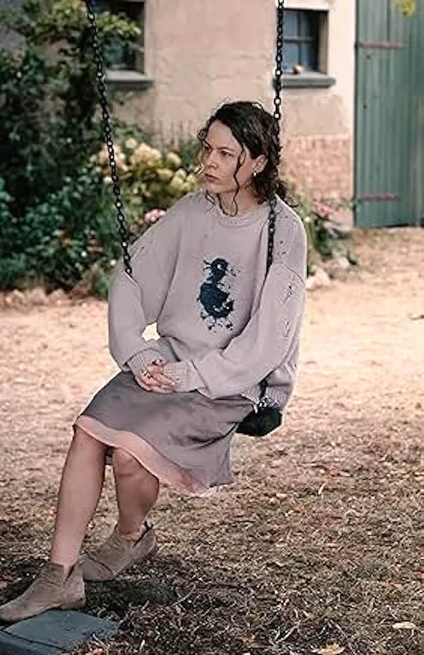A still from the film Mirrors No. 3. A white woman sits on a swing in a garden. She is wearing a grey jumper with a black duch silhouette on it.